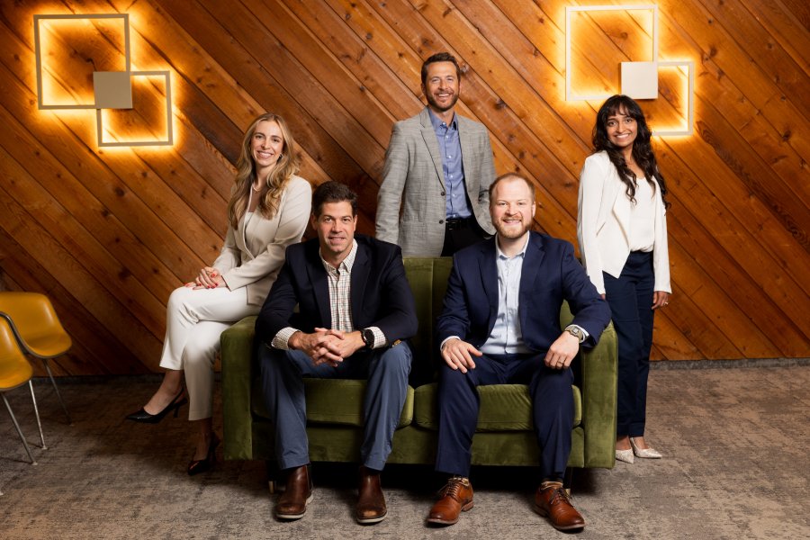20260521-restore0104-085A3929 | Restore Dental Arts Five professionals pose for a team photo in a wood-paneled office; two men sit on a green couch while three colleagues stand behind and to the sides, all smiling.