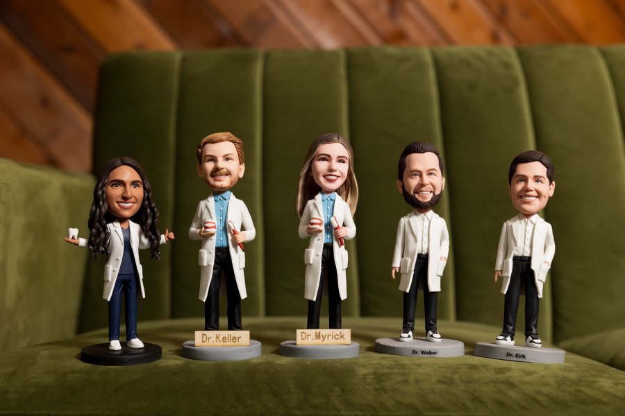 Five doctor bobbleheads in white lab coats stand on a green sofa; from left: unnamed, Dr. Keller, Dr. Myrick, Dr. Weber, Dr. Kirk on their bases.