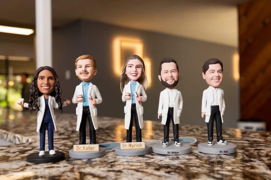 Five bobblehead doctors in white lab coats stand on a marble countertop; labels read Dr. Keller, Dr. Myrick, Dr. Weber, and Dr. Kirk (one figure unnamed).