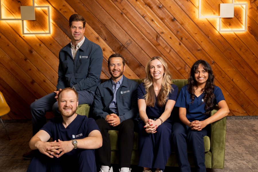 Five healthcare professionals sit and pose on a green couch in a wood-paneled room with modern square wall lights, wearing navy scrubs and labeled jackets for a team photo.