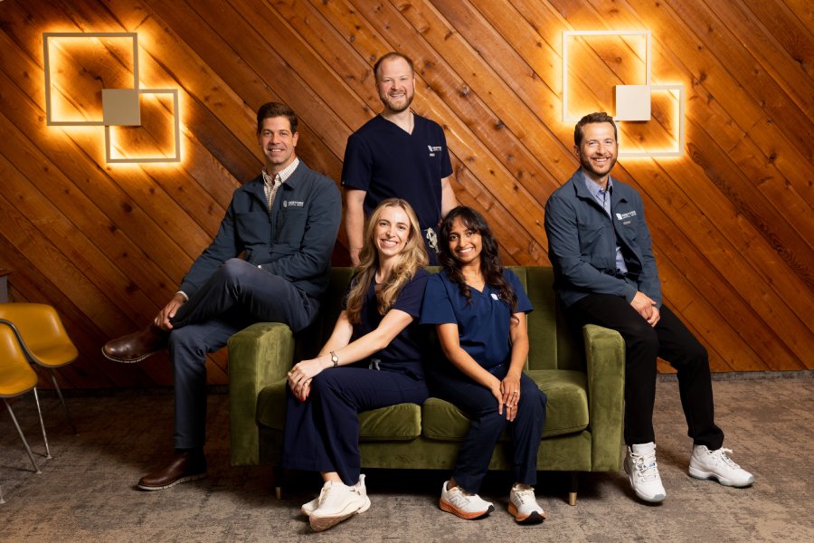 20260521-restore0126-085A4008 | Restore Dental Arts Group photo of healthcare professionals posing on a green couch in a wood-paneled room with geometric wall lights, smiling at the camera.
