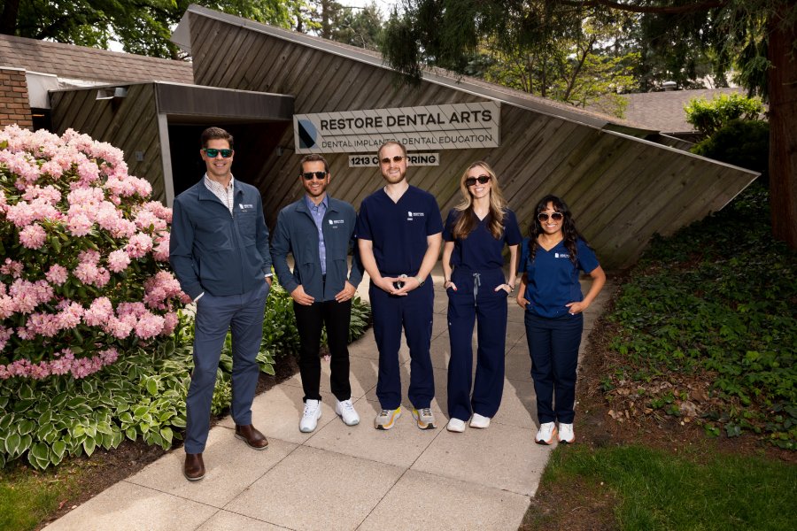 Five dental professionals pose outside Restore Dental Arts, wearing scrubs and casual jackets, smiling at the camera.