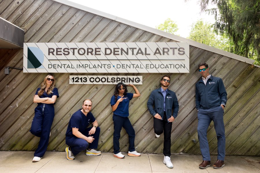 Six dental professionals in scrubs and jackets pose outside a wood-clad clinic wall beneath a sign that reads Restore Dental Arts and 1213 Coolspring.