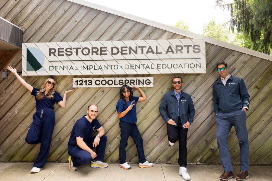 Five dental team members in scrubs posing outside a wooden wall sign that reads Restore Dental Arts, Dental Implants, Dental Education, 1213 Coolspring.
