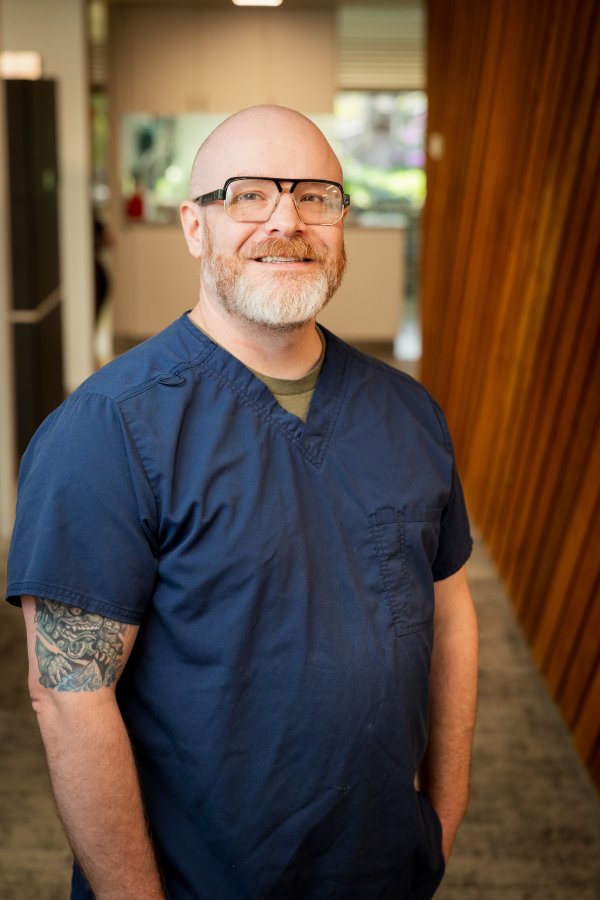 Smiling male healthcare worker with glasses in navy scrubs standing in a clinic hallway.
