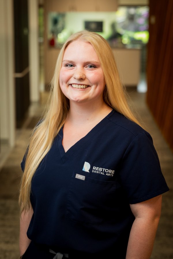 Dental staff member in navy scrubs with 'Restore Dental Arts' logo, smiling in a clinic hallway.