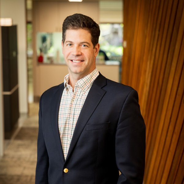 Dr Kirk | Restore Dental Arts Professional headshot of a smiling man in a navy blazer and plaid shirt, standing in a modern office hallway.