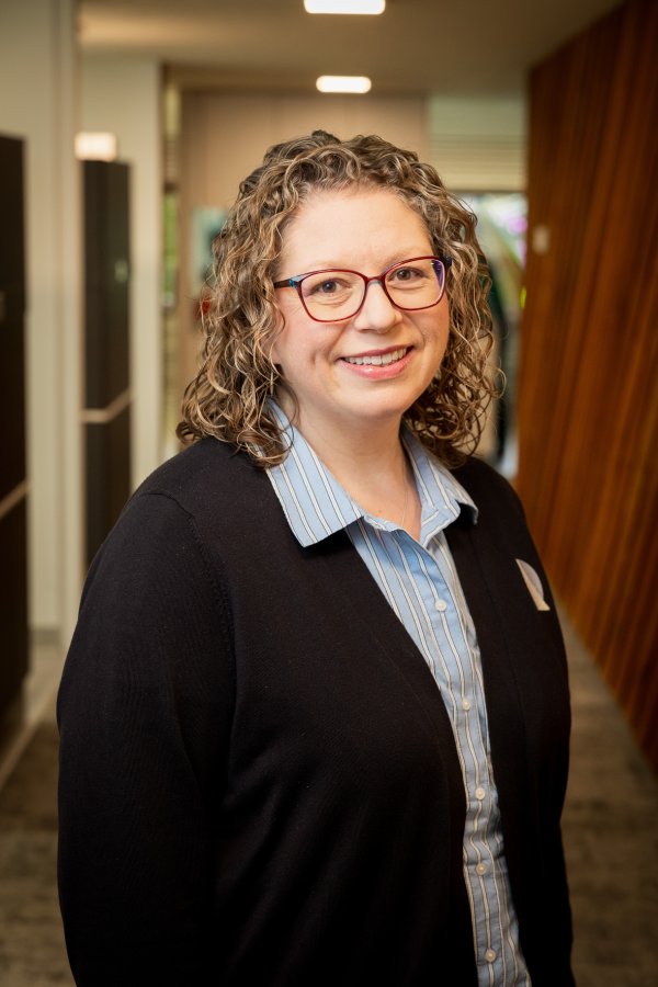 Smiling woman with curly hair and red glasses, wearing a blue striped shirt and black cardigan in an office hallway.