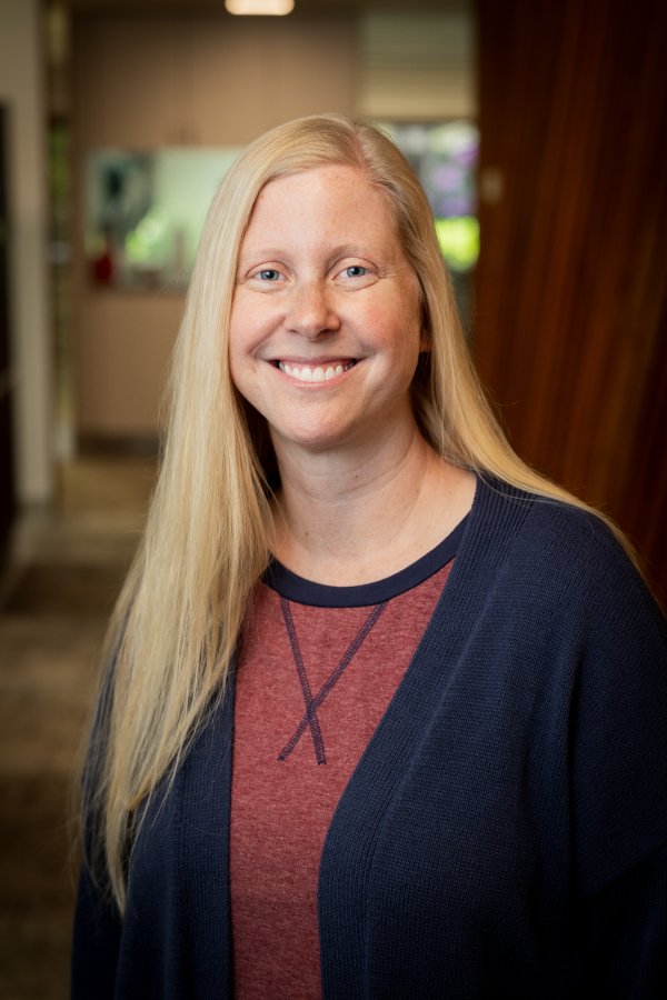 Head-and-shoulders portrait of a smiling woman with long blonde hair, wearing a maroon shirt and navy cardigan in a bright indoor setting.