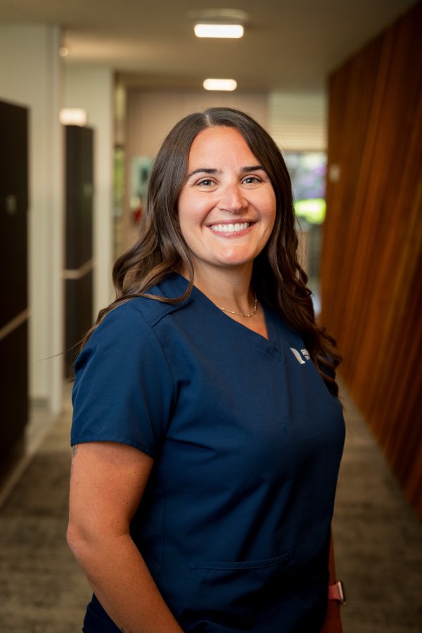Smiling female healthcare worker in navy scrubs standing in a clinic hallway.