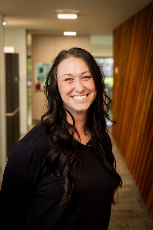 Smiling woman with long dark hair wearing a black top in a bright hallway.
