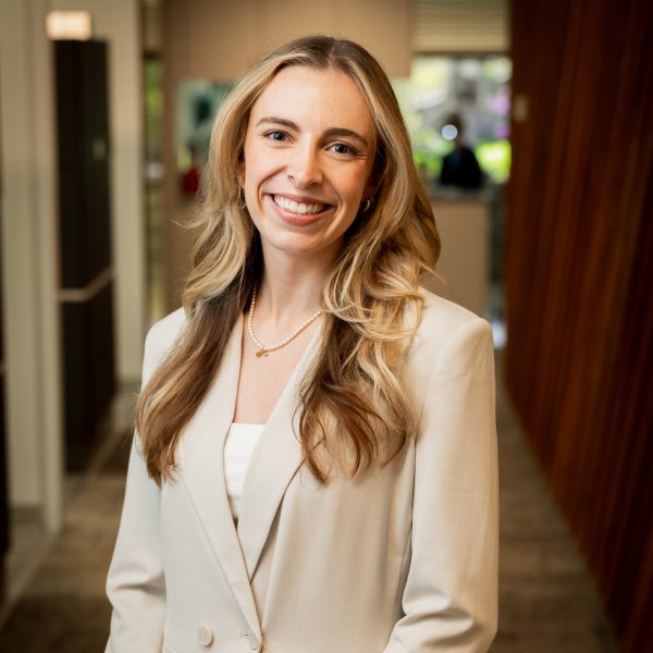 Professional portrait of a smiling woman in a light blazer standing in an office hallway.