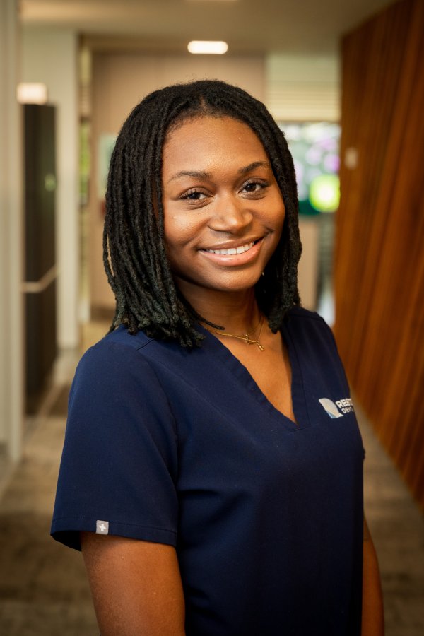 Friendly healthcare professional in navy scrubs standing in a clinic hallway, smiling at the camera with a name/logo on the uniform.