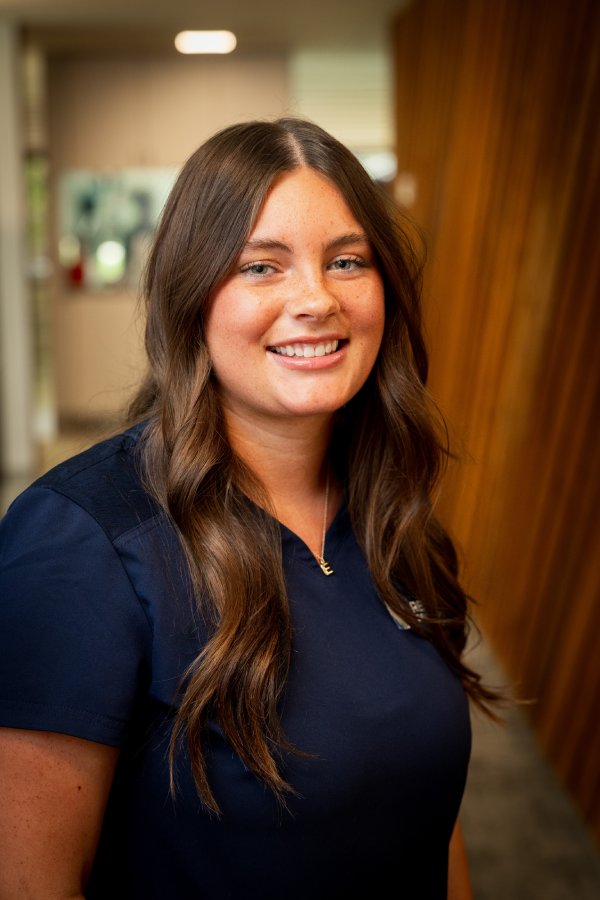 Smiling young woman in dark blue scrubs standing in a hallway, facing the camera.