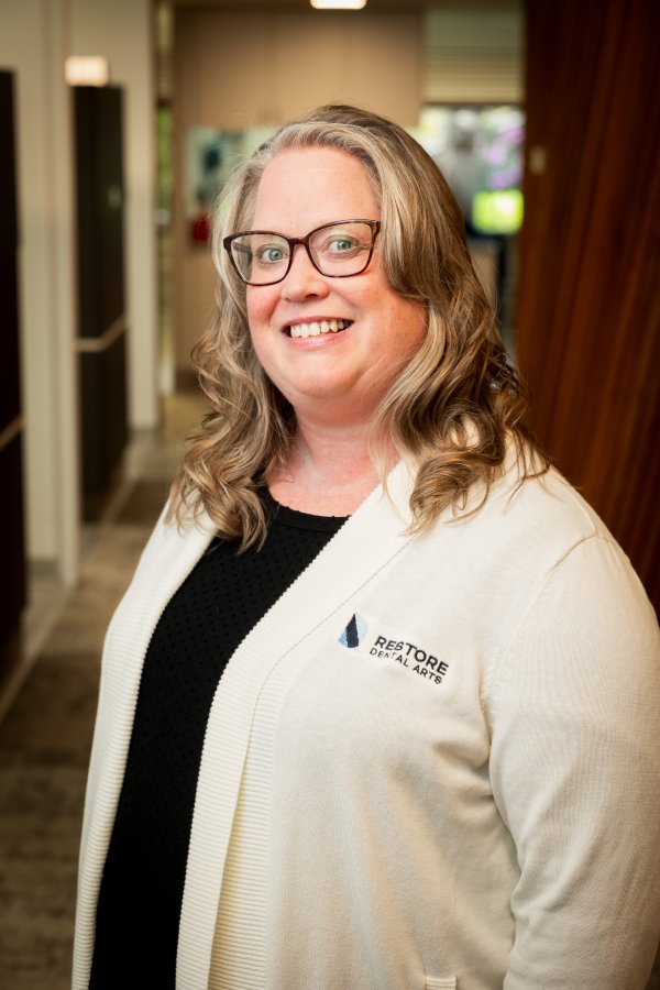 Smiling woman with glasses in a cream cardigan inside a dental office, wearing a Restore Dental Arts logo.