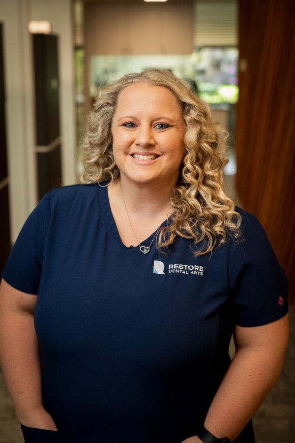 Smiling dental professional in navy scrubs with Restore Dental Arts logo, standing in a clinic hallway.