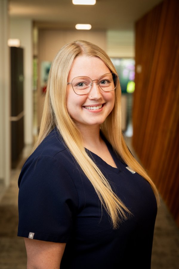 Smiling young woman with long blonde hair and round glasses wearing navy scrubs in a clinic hallway