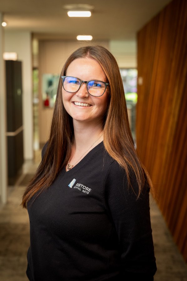 Smiling woman with long brown hair and blue glasses in a hallway, wearing a black shirt with a small logo on the chest (Restore Digital Arts).