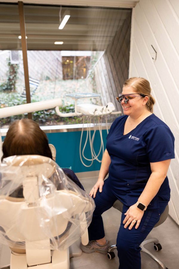 Dental hygienist in navy scrubs and safety glasses sits beside a patient in a dental chair, dental equipment visible in the background.