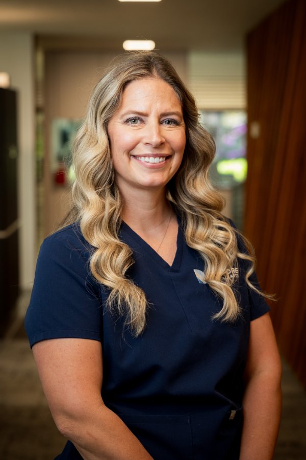Smiling woman with long wavy blonde hair in navy scrubs standing in a clinic hallway
