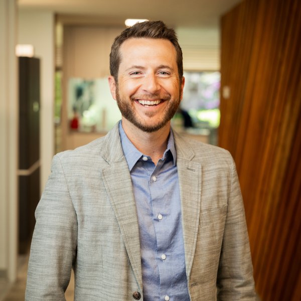 Professional headshot of a smiling man in a light gray blazer and blue shirt, standing in a modern office.