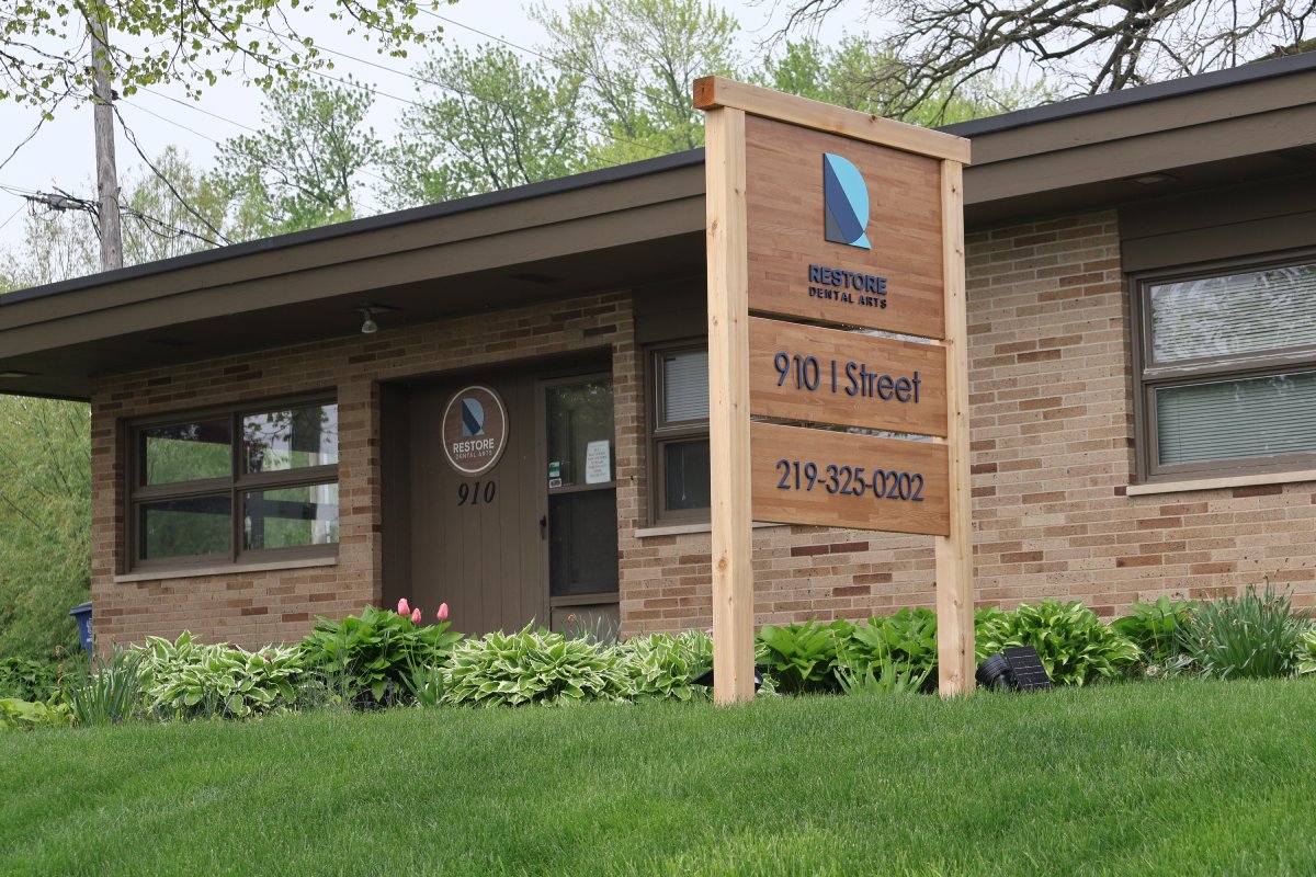 Brick dental clinic exterior with a tall wooden sign reading Restore Dental Arts, 910 I Street, phone 219-325-0202.