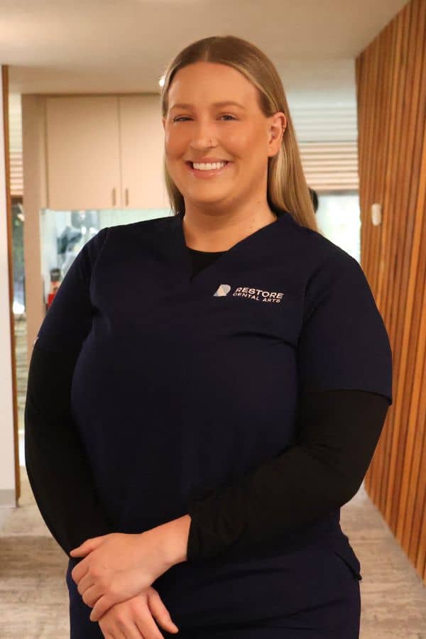 Smiling woman in navy scrubs with the 'Restore Dental Arts' logo in a clinic hallway.