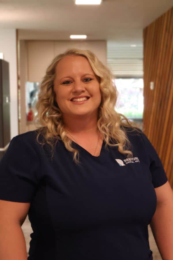 Smiling blonde dental assistant in navy scrubs stands in a clinic hallway.