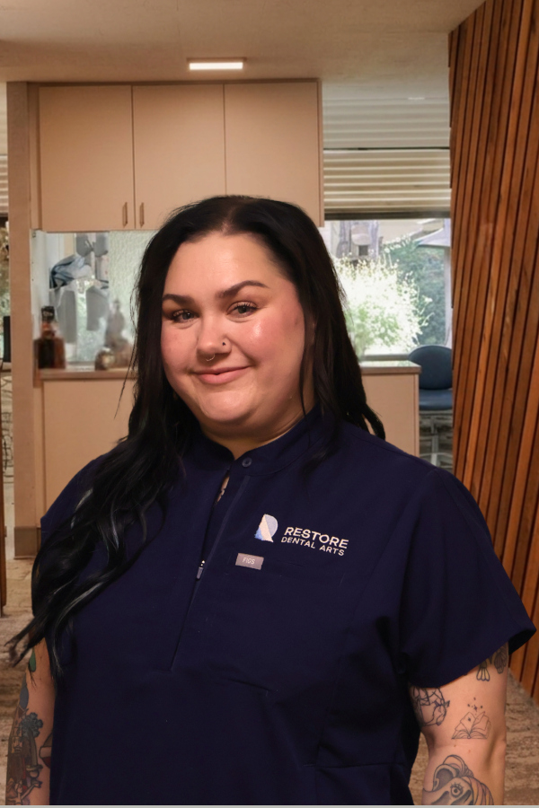 Smiling woman in navy dental scrubs standing in a clinic hallway with cabinets in the background, logo visible on her shirt