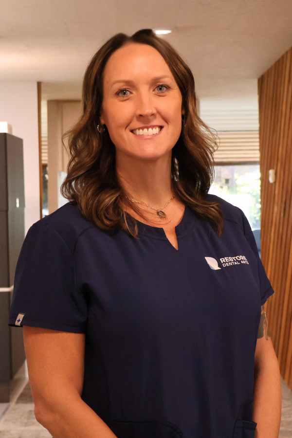Smiling woman in navy scrubs standing in a dental office, logo reading 'Restore Dental Arts' on her shirt