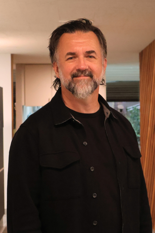 Smiling man with dark hair and gray beard wearing a black jacket indoors.
